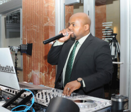 A man in a suit DJs at an event, speaking into a mic behind a mixer and @djbabyface504 laptop—Louisiana wedding inspiration.