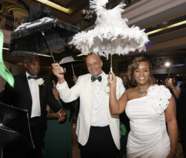 Man and woman in white formal wear smile with feathered umbrellas at an elegant New Orleans wedding celebration indoors.