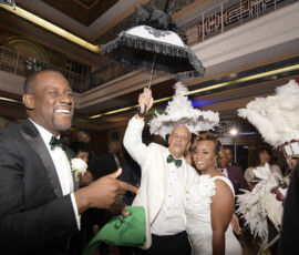 Three people in formal attire celebrate indoors; one holds a black-and-white umbrella amid feathered hats and Louisiana wedding style.