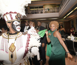 A guest in a feathered costume dances beside a woman in green as others celebrate in a festive, New Orleans-style ballroom.