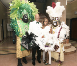 Two people in feathered costumes pose with a bride and groom on a marble floor near stairs at a vibrant New Orleans wedding.