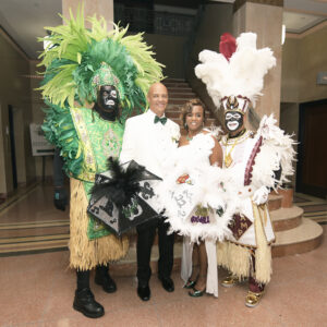 Two people in feathered costumes pose with a bride and groom on a marble floor near stairs at a vibrant New Orleans wedding.