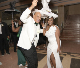 Two people in formal white dance with feathered umbrellas at an indoor event, evoking a lively New Orleans wedding.