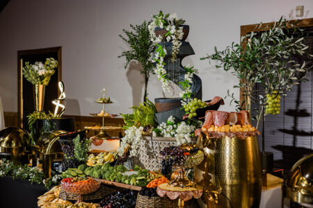 Elegant buffet table with fruits, cheese, meats, and bread, accented by flowers and a tiered water fountain centerpiece.