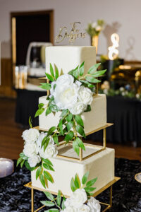 Elegant three-tier white wedding cake with gold stands, white roses, and “The Driggers” topper on black tablecloth.