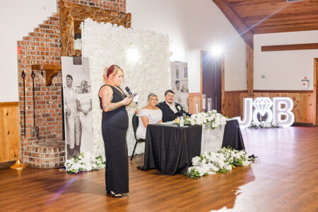 Woman in black dress gives speech beside newlyweds at Louisiana wedding with white flowers, brick fireplace, and J♥B initials.