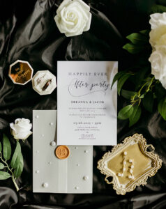 Elegant wedding invitation flat lay with green envelope, wax seal, gold tray, pearl jewelry, ring box, and white roses.