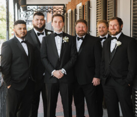 Six men in black tuxedos pose on a brick walkway outside a building with white doors—classic Louisiana wedding inspiration.