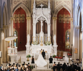 Bride and groom at altar in ornate Gulf South church with tall arches, red walls, and guests seated in pews.