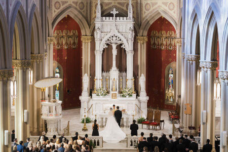 Bride and groom at altar in ornate Gulf South church with tall arches, red walls, and guests seated in pews.