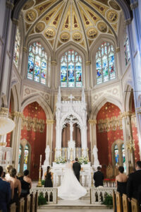 Bride and groom at a grand Louisiana church altar with stained glass, gold accents, and guests watching from pews.