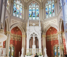 Bride and groom at a grand Louisiana church altar with stained glass, gold accents, and guests watching from pews.