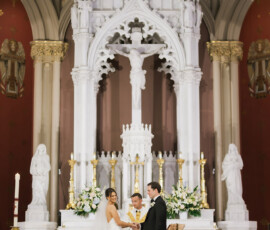 Bride and groom hold hands at a grand church altar, priest officiating amid ornate white and gold Gulf South wedding decor.