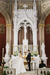 Bride and groom hold hands at a grand church altar, priest officiating amid ornate white and gold Gulf South wedding decor.