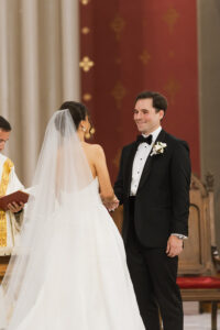 Bride and groom holding hands, smiling at the altar with a priest during their Louisiana wedding ceremony.