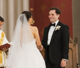 Bride and groom holding hands, smiling at the altar with a priest during their Louisiana wedding ceremony.