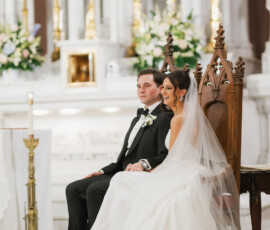 Bride and groom in classic attire sit on ornate chairs at a church altar, surrounded by flowers and candles.
