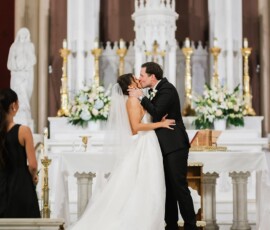 Bride and groom kiss at a Louisiana church altar, surrounded by white flowers, statues, and a nearby bridesmaid in black.