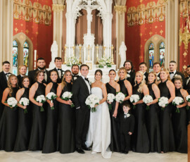 Bride and groom with wedding party in black attire pose in a church, holding white bouquets amid elegant details.