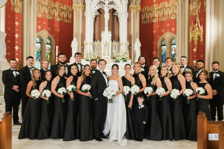 Bride and groom with wedding party in black attire pose in a church, holding white bouquets amid elegant details.