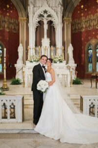 Bride and groom in wedding attire embrace, smiling before a decorated church altar with white flowers and statues.