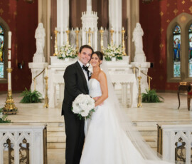 Bride and groom in wedding attire embrace, smiling before a decorated church altar with white flowers and statues.