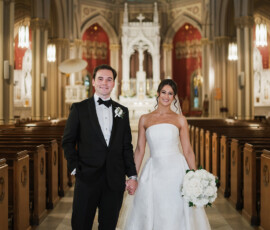 Bride in strapless white gown and groom in black tuxedo smile inside ornate church with stained glass windows.