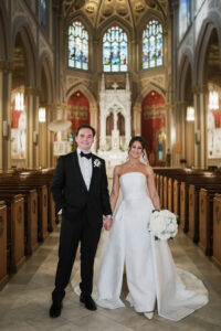 Bride in strapless white gown and groom in black tuxedo smile inside ornate church with stained glass windows.