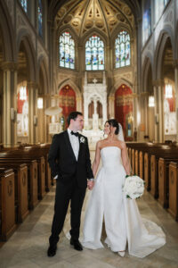 Bride in strapless gown with white flowers and groom in black tuxedo hold hands in grand New Orleans church wedding.