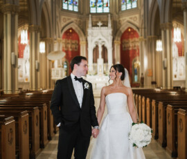 Bride in strapless gown with white flowers and groom in black tuxedo hold hands in grand New Orleans church wedding.