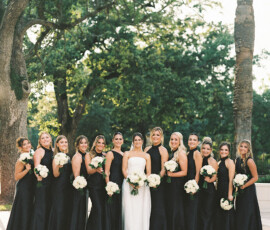 Bride in white with bridesmaids in black dresses holding white bouquets, outdoors with trees—Gulf South wedding inspiration.