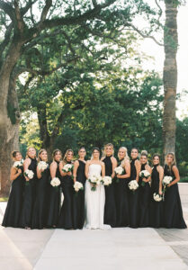 Bride in white with bridesmaids in black dresses holding white bouquets, outdoors with trees—Gulf South wedding inspiration.