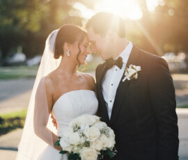 Bride with white bouquet and veil smiles at groom in tuxedo, sun shining behind, perfect for Gulf South wedding inspiration.