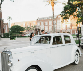 Classic white car parked on a sunny street, two people nearby, trees and historic brick New Orleans building in the background.