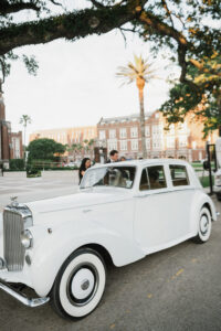 Classic white car parked on a sunny street, two people nearby, trees and historic brick New Orleans building in the background.