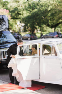 Man in suit assists woman in wedding dress from white classic car at Louisiana wedding, with greenery and parked cars behind.