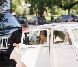 Man in suit assists woman in wedding dress from white classic car at Louisiana wedding, with greenery and parked cars behind.
