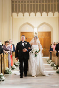 Bride in white gown and veil walks down aisle with man in tuxedo in decorated church, classic New Orleans wedding scene.