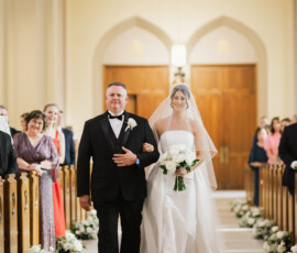 Bride in white gown and veil walks down aisle with man in tuxedo in decorated church, classic New Orleans wedding scene.