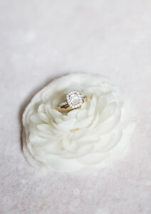 Diamond halo engagement ring on layered white flower atop a pale pink surface—perfect for a romantic Louisiana wedding.