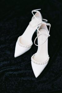 White pointed-toe heels with ankle straps and pearl accents on a textured black fabric, ideal for a New Orleans wedding.