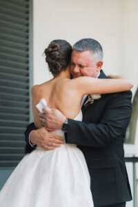 Woman in white dress hugs older man in suit near window, sharing an emotional moment before the wedding ceremony.