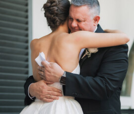 Woman in white dress hugs older man in suit near window, sharing an emotional moment before the wedding ceremony.