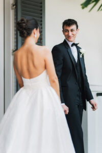 Bride in white gown and groom in tuxedo smile outside near white walls and green shutters at a New Orleans wedding.