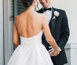 Bride in strapless white gown faces groom in black tuxedo by window with green shutters at New Orleans wedding ceremony.