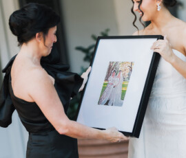 Two women, one in white and one in black dresses, smile holding a framed photo outside at a possible Louisiana wedding.