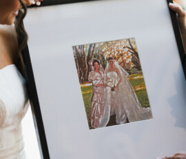 Woman in white dress holds framed photo of two brides outdoors; another person’s hands and arms also hold the frame.