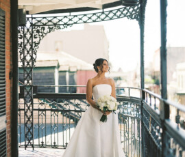 Bride in strapless white gown with bouquet smiles on ornate sunlit balcony, elegant New Orleans wedding.