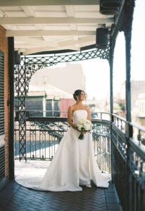 Bride in strapless white gown with bouquet smiles on ornate sunlit balcony, elegant New Orleans wedding.