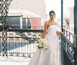 Bride in strapless white dress smiles on ornate balcony with bouquet; sunlight highlights Gulf South wedding elegance.
